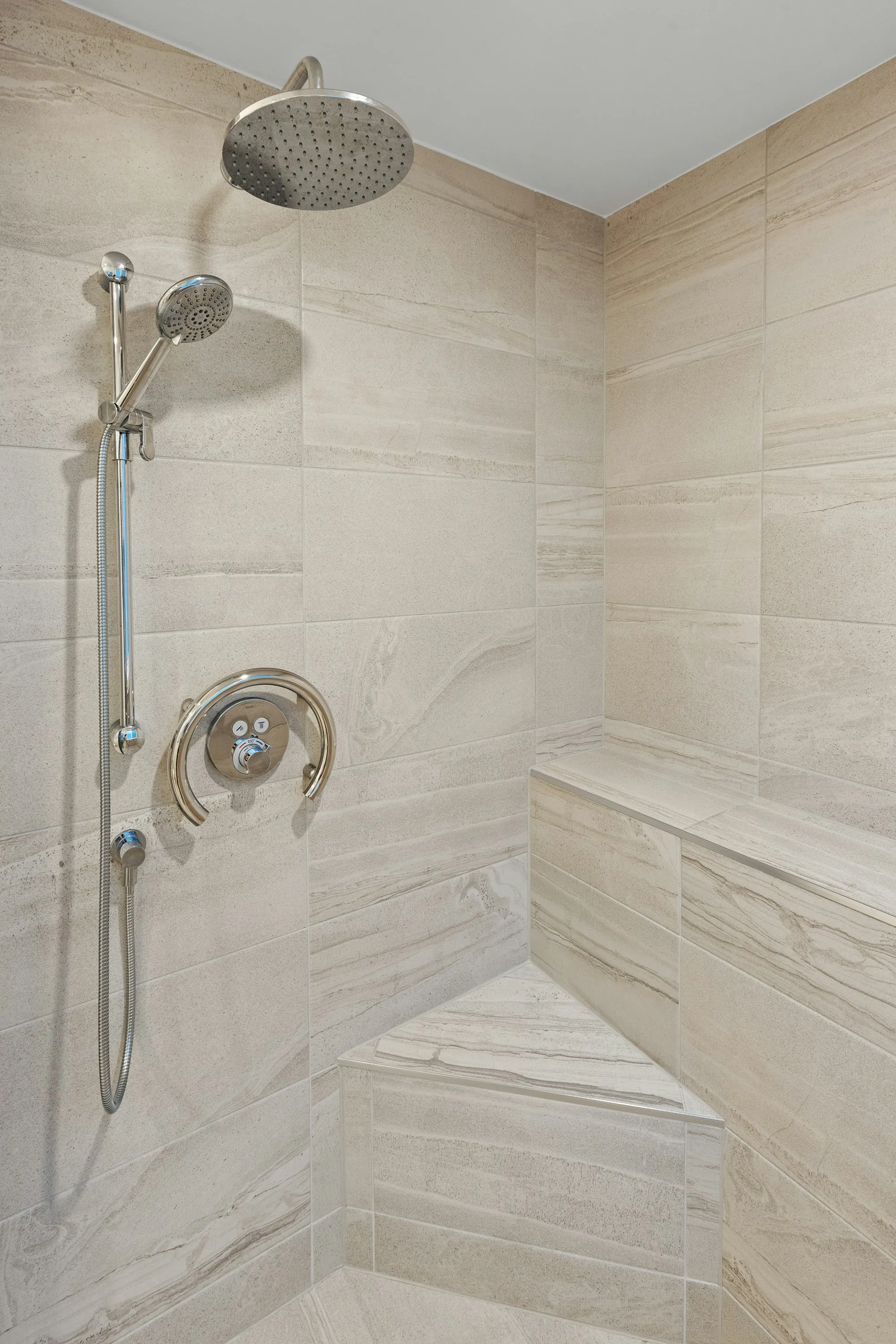 Walk-in shower with large stone-look tile, a corner bench and a handheld shower head